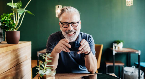 Mann mit grauen Haaren und weißer Brille hält Tasse in der Hand und sitzt vor grüner Wand