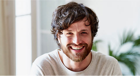 Nahaufnahme von dunkelhaarigem Mann mit weißem Oberteil, der in die Kamera lächelt