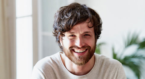 Mann mit dunklen Haaren in weißem Oberteil lacht in die Kamera