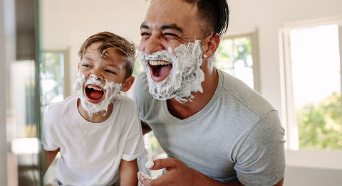Vater und Sohn stehen lachend mit Rasierschaum im Gesicht im Badezimmer