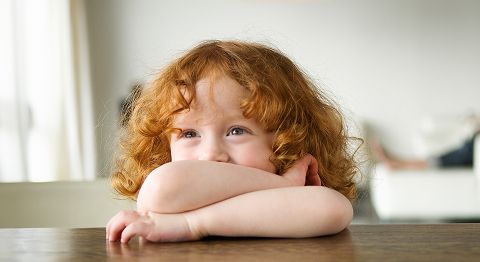Kind mit orangenen Haaren lehnt sich mit den Armen auf den Tisch und grinst
