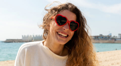 Frau mit Locken und Sonnenbrille in Herzform ist glücklich am Strand