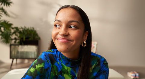 Frau mit dunkelbraunen Haaren und buntem Oberteil sitzt in ihrer Wohnung und lächelt in die Ferne