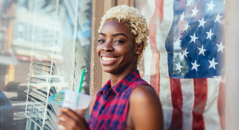 Frau mit blonden Locken in kurzen Karohemd steht vor einer USA-Flagge