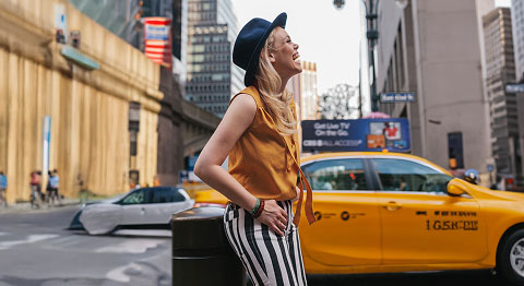 Blonde Frau mit Hut auf dem Kopf lacht in die Ferne und steht vor einem gelben Taxi