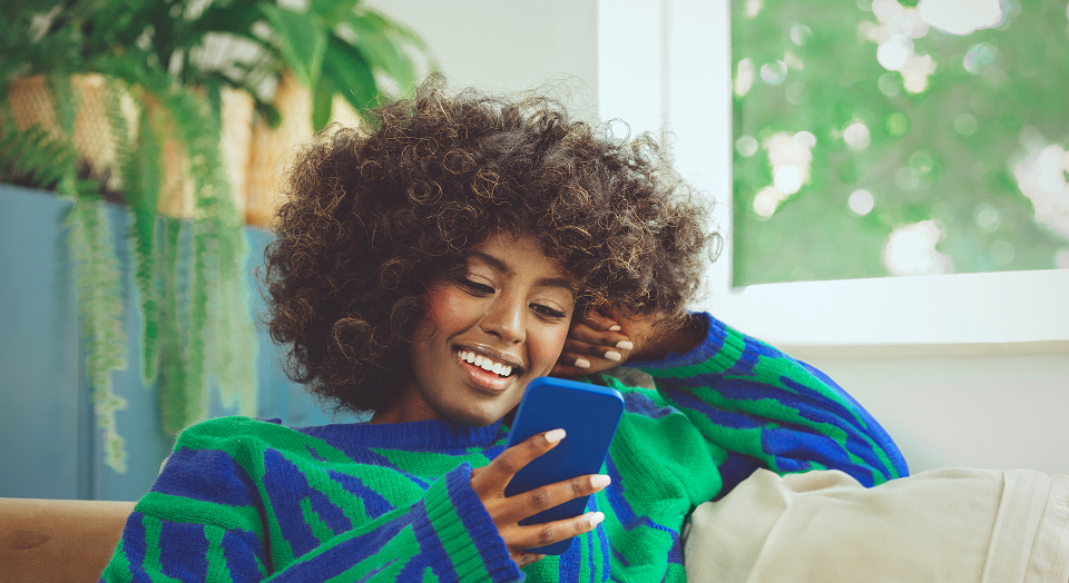 Lockige Frau in buntem Pullover sitzt auf einem Sofa und schaut in ihr Smartphone