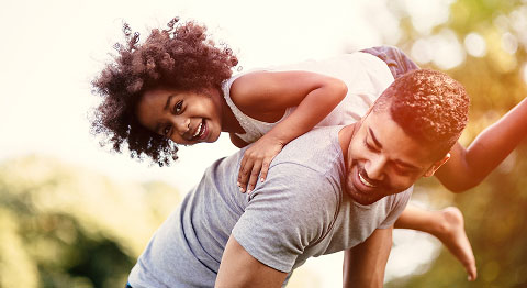 Vater hat Kind auf seinem Rücken und im Hintergrund sieht man grüne Bäume