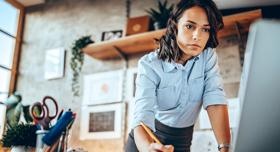 Braunhaarige Frau in Jeanshemd steht an ihrem Schreibtisch um Büro und schaut in ihren PC