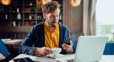 Mann mit braunen Haaren und Bart in einem Café, in der einen hält er eine Kaffeetasse, in der anderen ein Handy, vor ihm steht ein Laptop
