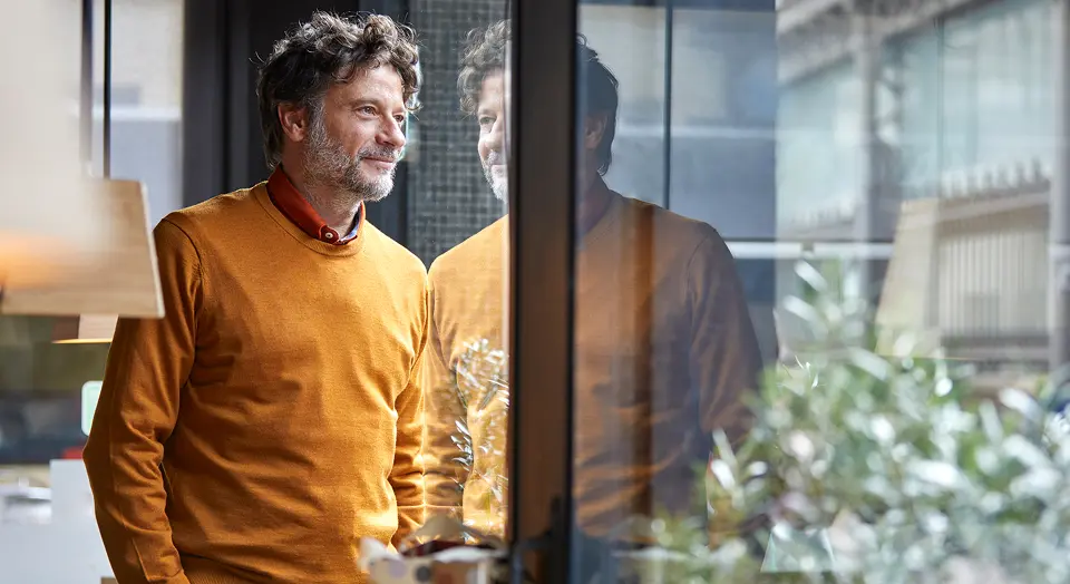 Mann in einem senfgelben Pullover mit grauem Bart und lockigen dunklen Haaren lehnt an einer Fensterscheibe und schaut hinaus.
