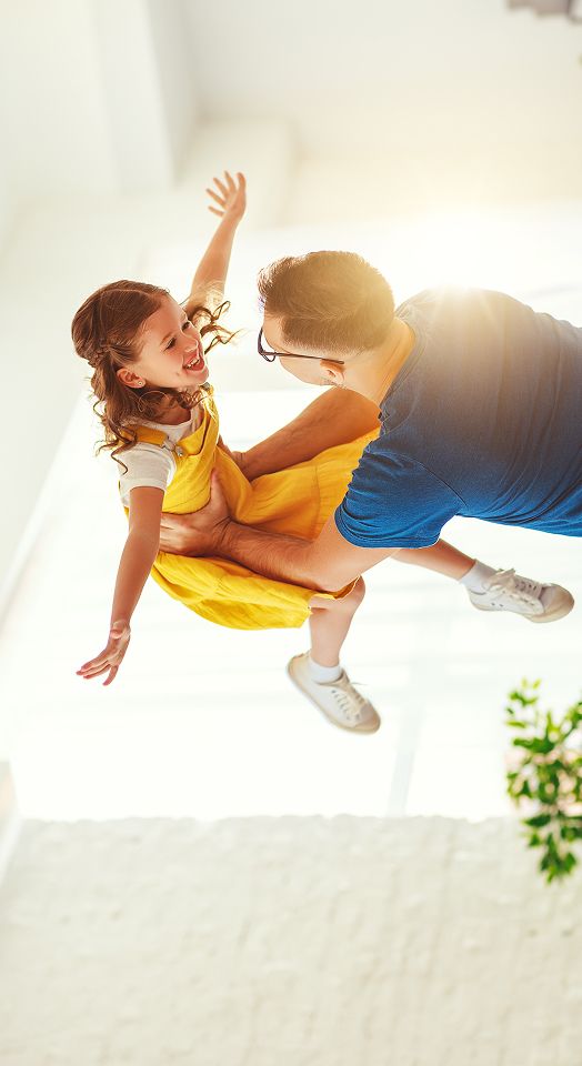 Vater in blauem Shirt und mit Brille wirft seine Tochter im gelben Kleid in die Luft und hält sie dabei fest