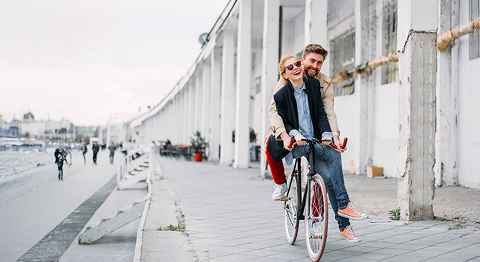 Frau mit Sonnenbrille und Mann mit braunen Haaren fahren zusammen auf dem Fahrrad und lachen
