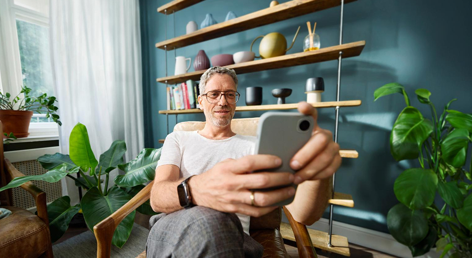 Mann mit Brille sitzt vor einem Regel auf einem Sessel und schaut in sein Smartphone