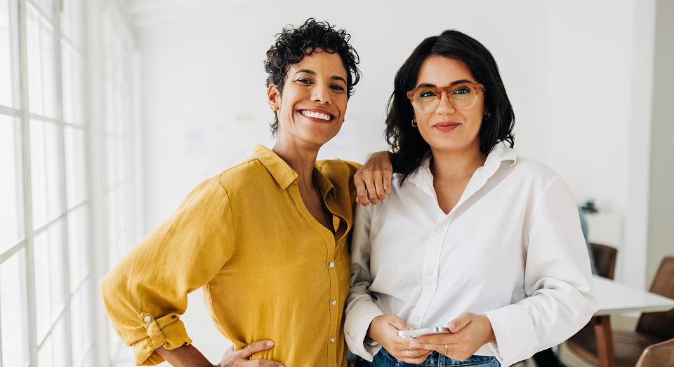 Zwei Frauen in Hemden lehnen sich aneinander und lächeln in die Kamera