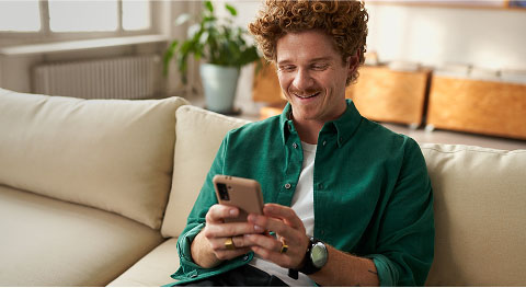 Mann mit orangenen Locken in grünem Hemd sitzt auf dem Sofa und schaut zufrieden in sein Smartphone