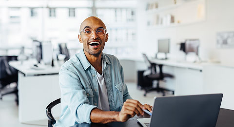 Mann mit Glatze mit Brille in Jeansjacke sitzt im Büro am Schreibtisch und lacht