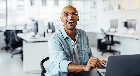Mann mit Glatze und Brille in Jeanshemd sitzt im Büro am Schreibtisch und lacht
