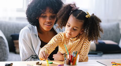 Frau mit Kind auf dem Schoss sitzt am Tisch und schaut dem Kind beim Malen zu, im Vordergrund ein Glas mit Stiften