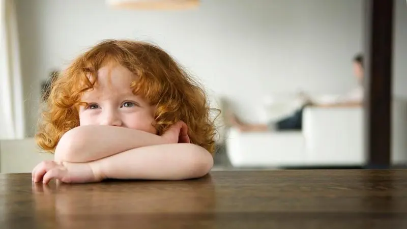 Mädchen mit roten Haaren stützt sich mit verschränkten Armen auf einen Tisch, im Hintergrund eine unscharfe person auf einem Sofa