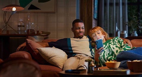 Mann und Frau sitzen aneinander gelehnt auf dem Sofa und schauen gemeinsam in ein blaues Smartphone