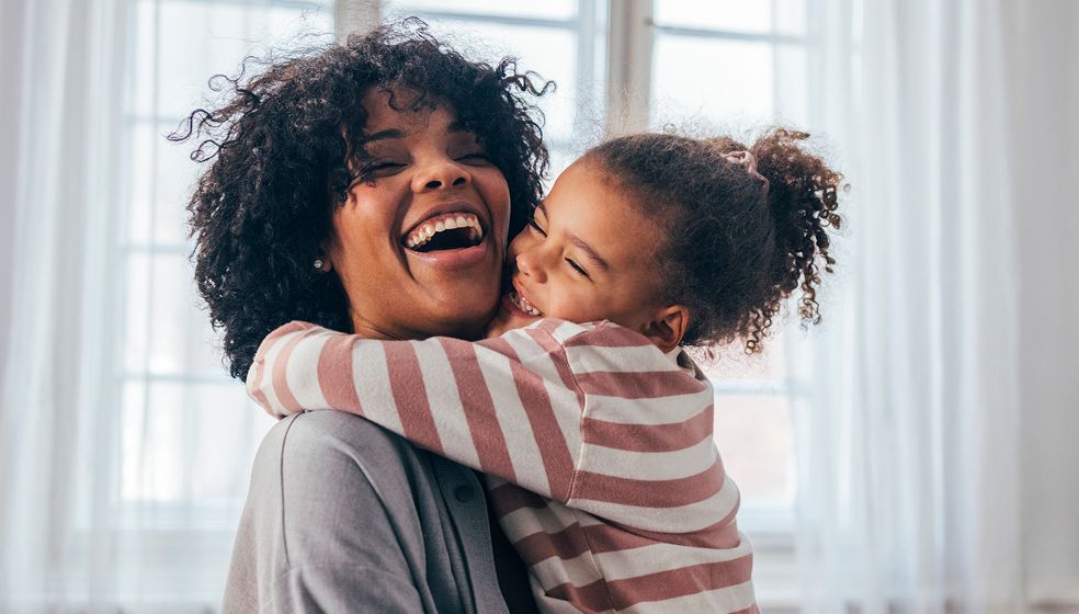 Kind umarmt Frau und beide lachen dabei, im Hintergrund ist ein Fenster