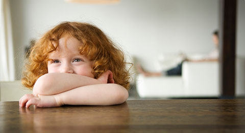 Kind mit orangenen Locken stützt sich auf seinen Händen am Tisch ab