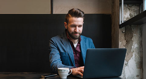 Dunkelhaariger Mann in blauem Sakko sitzt an seinem Laptop neben einer Tasse