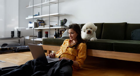 Dunkelhaarige Frau sitzt vor ihrem Sofa am Laptop und ihr kleiner weißer Hund liegt hinter ihr auf dem Sofa