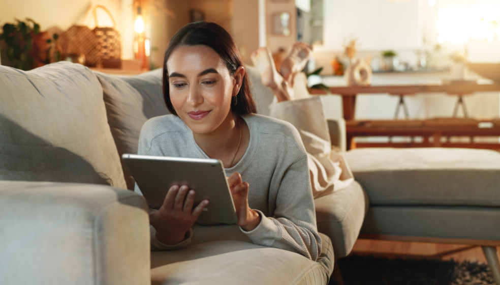 Frau liegt entspannt auf dem Sofa und nutzt ein Tablet in einem hellen Wohnzimmer