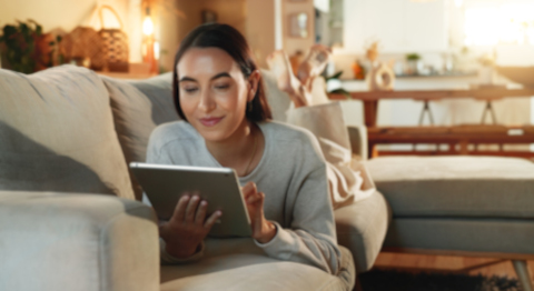 Frau liegt entspannt auf dem Sofa und nutzt ein Tablet in einem hellen Wohnzimmer