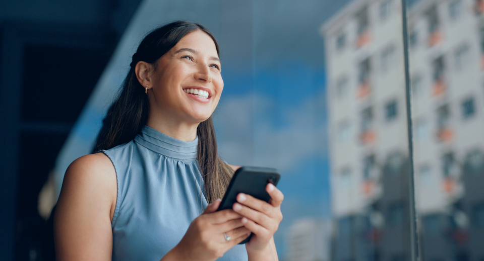 Frau mit schwarzen langen Haaren steht mit Smartphone in der Hand vor einer blauen Glaswand und lächelt
