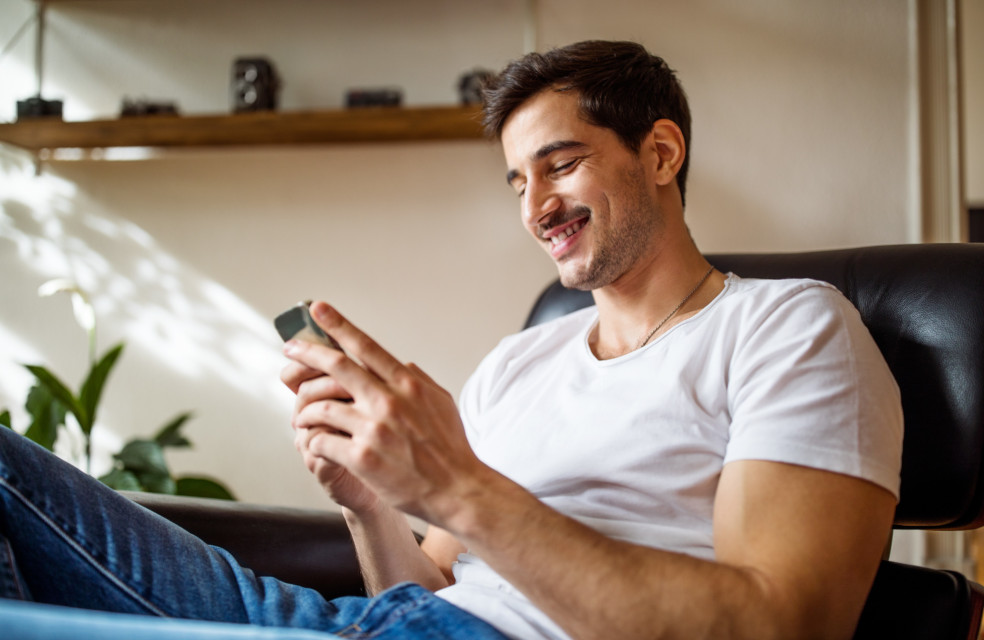 Mann mit kurzen braunen Haaren sitzt auf Sofa, lacht und schaut in sein Handy