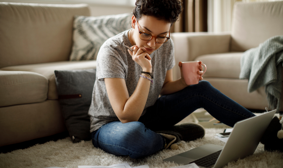 Frau mit kurzen braunen Haaaren und Brille sitzt auf dem Boden vorm Sofa und schaut auf Laptop