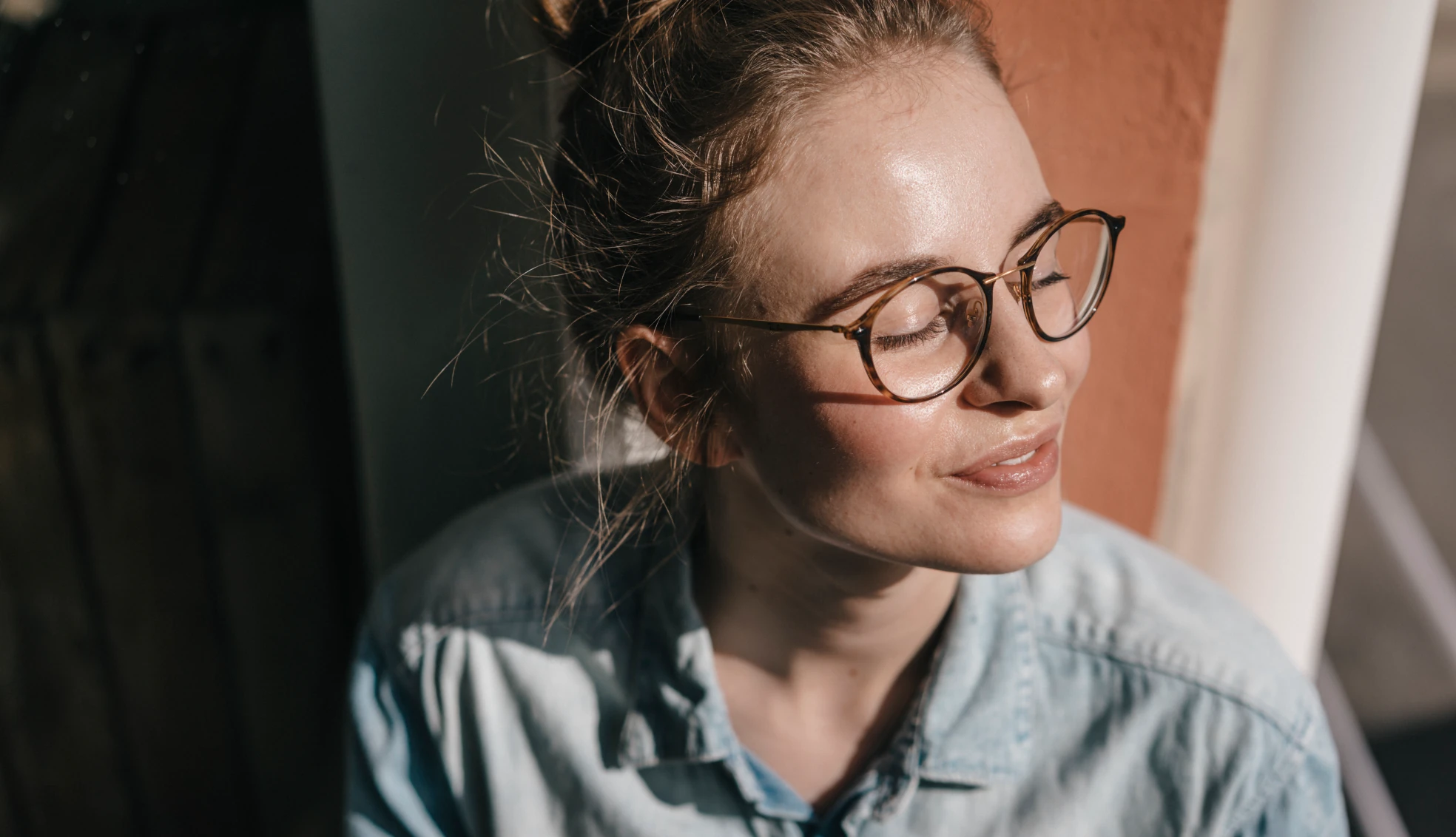 Frau mit Brille sitzt auf dem Boden vor dem Fenster mit geschlossenen Augen und genießt die Sonne
