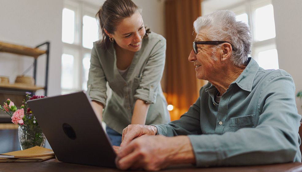 Das Bild zeigt zwei Personen in einem hellen Raum. Eine Person sitzt an einem Tisch und bedient einen Laptop, während die andere Person stehend daneben Unterstützung leistet. Auf dem Tisch liegen ein Notizbuch und ein Blumenstrauß. Im Hintergrund sind Fenster mit Tageslicht zu sehen.