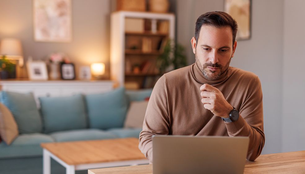 Das Bild zeigt eine Person in einem Wohnzimmer, die an einem Tisch vor einem Laptop sitzt. Im Hintergrund sind ein Sofa, ein Regal mit Büchern und Dekoration sowie weitere Möbel und Lichtquellen zu sehen. Die Person trägt einen braunen Pullover und eine Armbanduhr.