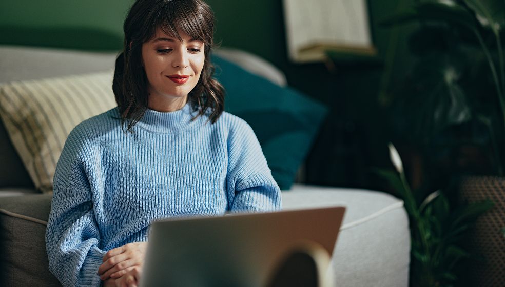 Eine Person sitzt auf einem Sofa vor einem geöffneten Laptop. Im Hintergrund sind Kissen, eine Pflanze und ein Bild zu sehen. Die Person trägt einen hellblauen Strickpullover.