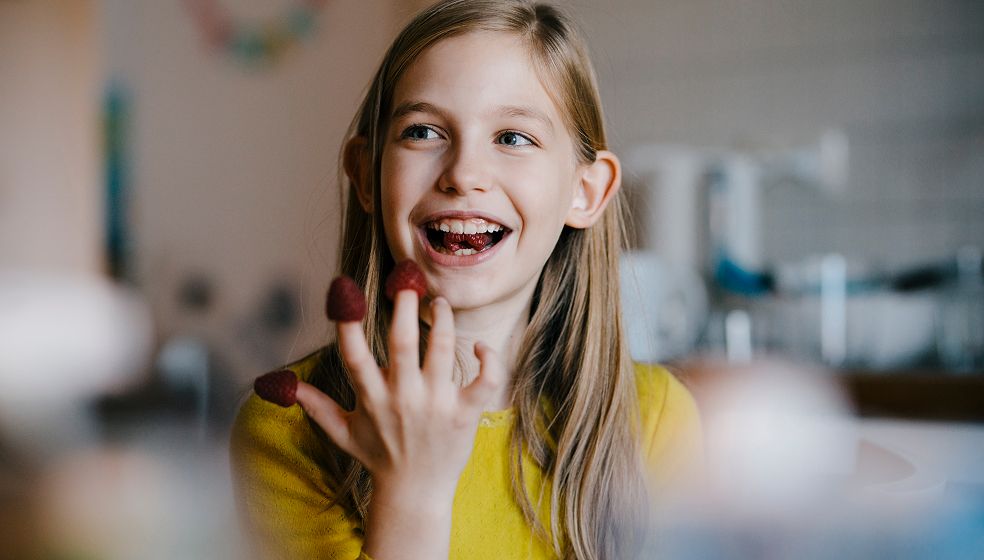 Mädchen in gelbem Pullover hat Himbeeren auf ihre Finger gesteckt und isst diese nach und nach
