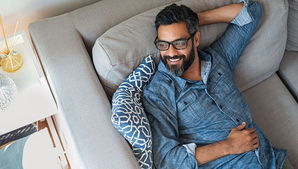 Ein Mann liegt entspannt auf einem hellgrauen Sofa und trägt ein blaues Hemd. Sein Kopf liegt auf einem blauen Kissen mit weißem Muster. Neben dem Sofa steht ein weißer Beistelltisch mit einer dekorativen Duftflasche und einer kleinen Lampe.