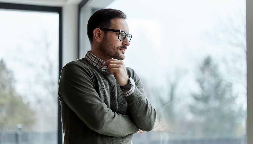 Das Bild zeigt eine Person mit kurzem dunklem Haar und Bart, die vor einem großen Fenster steht. Die Person trägt eine Brille, ein kariertes Hemd und einen grauen Pullover. Sie steht mit verschränkten Armen und denkt nach, wobei eine Hand das Kinn berührt. Im Hintergrund sind verschwommene Bäume und ein heller Himmel zu erkennen.