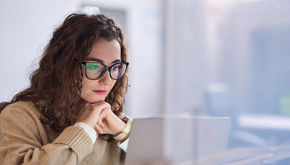 Eine Person mit lockigen braunen Haaren und Brille sitzt an einem Schreibtisch und arbeitet an einem Laptop. Sie trägt einen beigen Pullover und eine Armbanduhr. Die Umgebung wirkt wie ein Büro mit heller, unscharfer Einrichtung im Hintergrund.