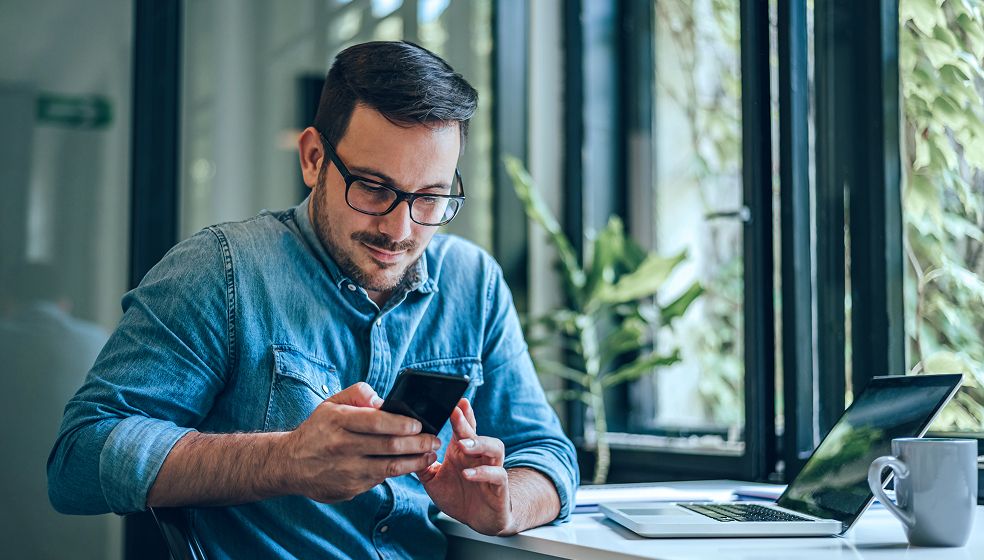 Ein Mann sitzt an einem Schreibtisch mit einem Laptop, einer Tasse und einem Smartphone in der Hand. Im Hintergrund sind große Fenster, durch die man Pflanzen sieht.