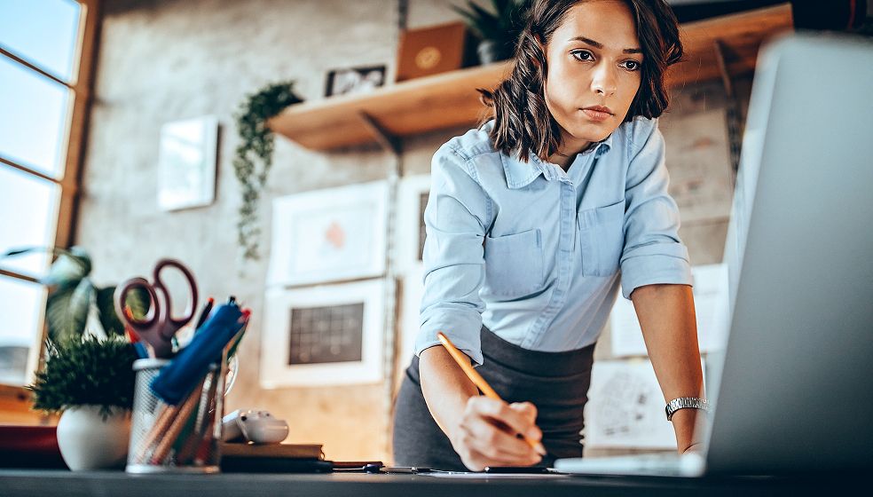 Dunkelhaarige Frau in hellblauem Hemd beugt sich unter an ihren PC auf dem Schreibtisch