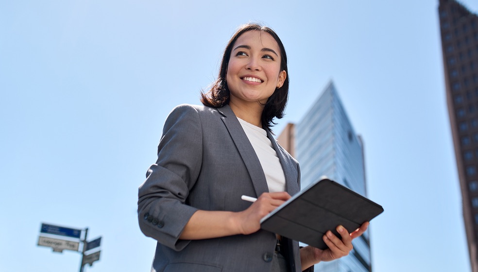 Frau  mit dunklen, halblangen Haaren im grauen Blazer steht vor zwei Hochhäusern,, hält ein Tablet in der Hand und blickt lächelnd zur Seite