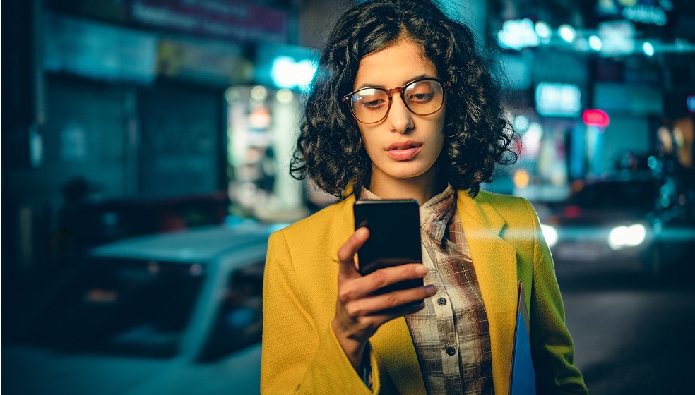 Frau mit braunen lockigen Haaren und Brille im gelben Blazer hält ihr Smartphone hoch und blickt gespannt auf den Bildschirm