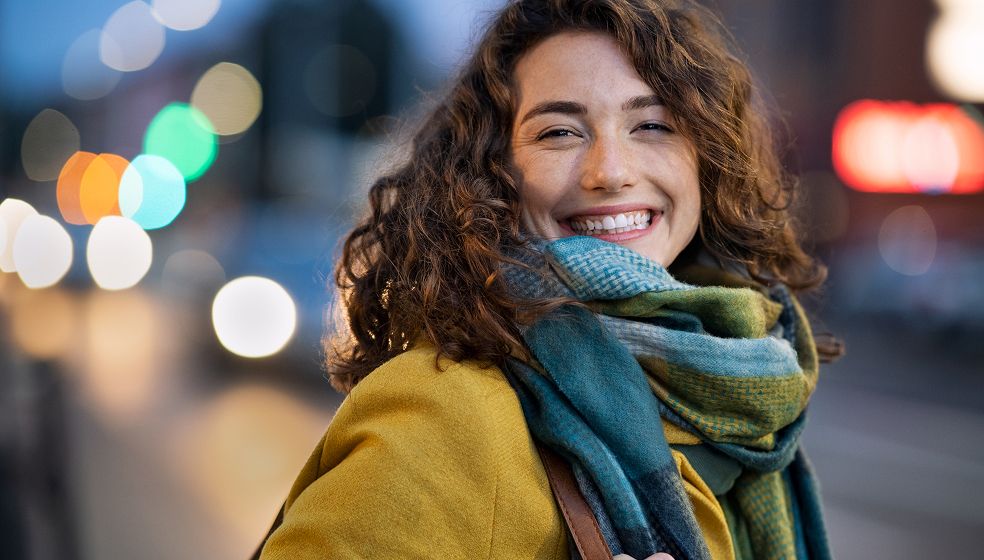 Das Bild zeigt eine Person mit lockigen, braunen Haaren, die einen gelben Mantel und einen grün-blauen Schal trägt. Die Person steht im Freien, vermutlich an einer Straße in einer Stadt bei Dämmerung oder Abend. Im Hintergrund sind unscharfe, leuchtende Lichter von Autos und Straßenbeleuchtung zu sehen.