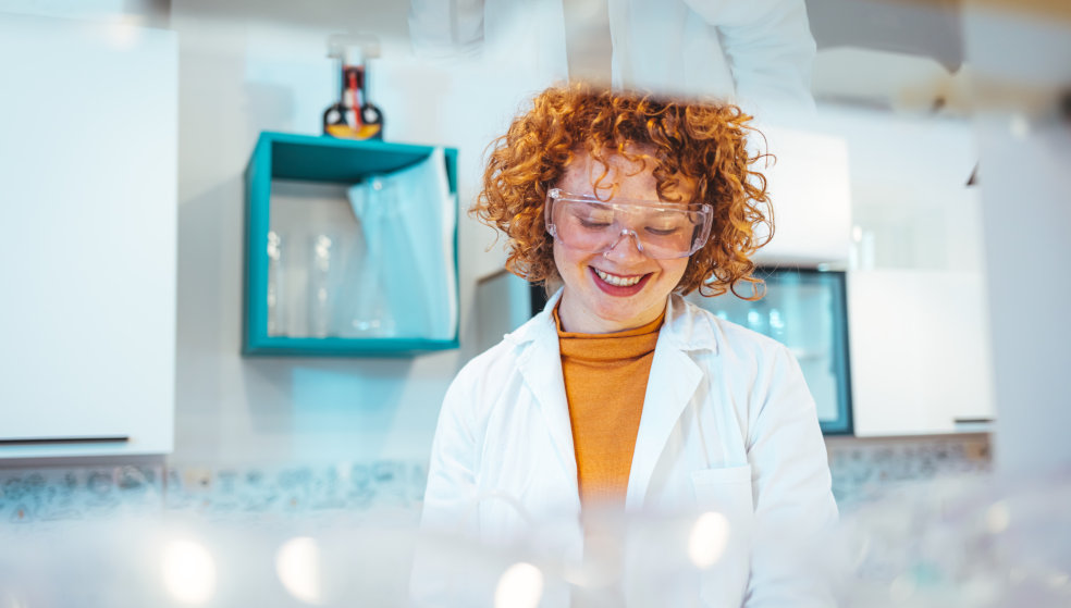 Frau mit orangenen Locken steht mit Schutzbrille und Kittel in einem Labor