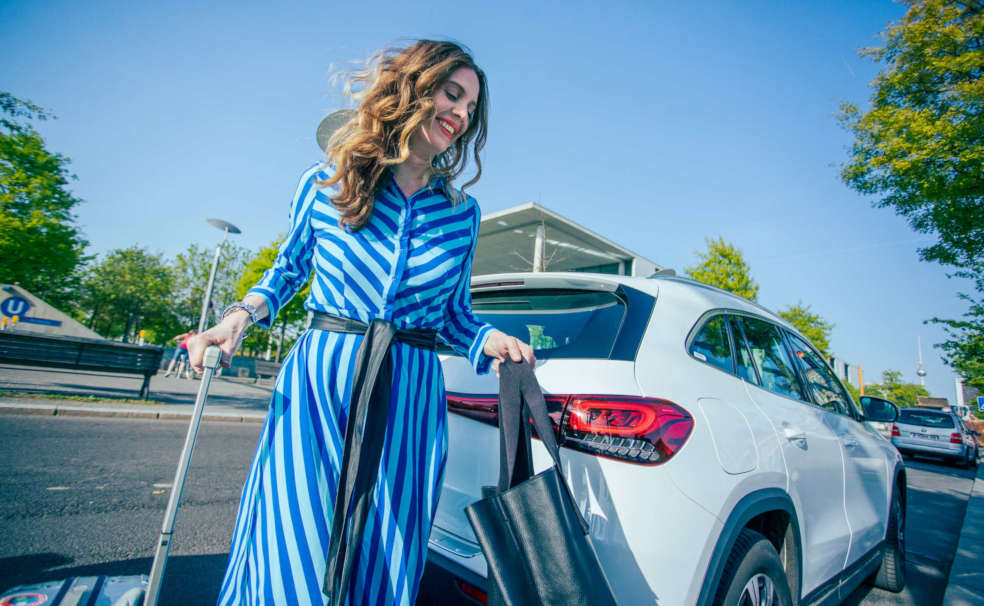 Lockige Frau in blau weiß gestreiftem Kleid läuft hinter einem weißen E-Auto