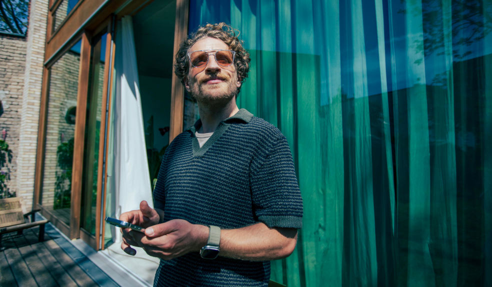 Mann mit Locken und Sonnenbrille steht vor einen Haus mit bodentiefen Fenster und schaut nach oben