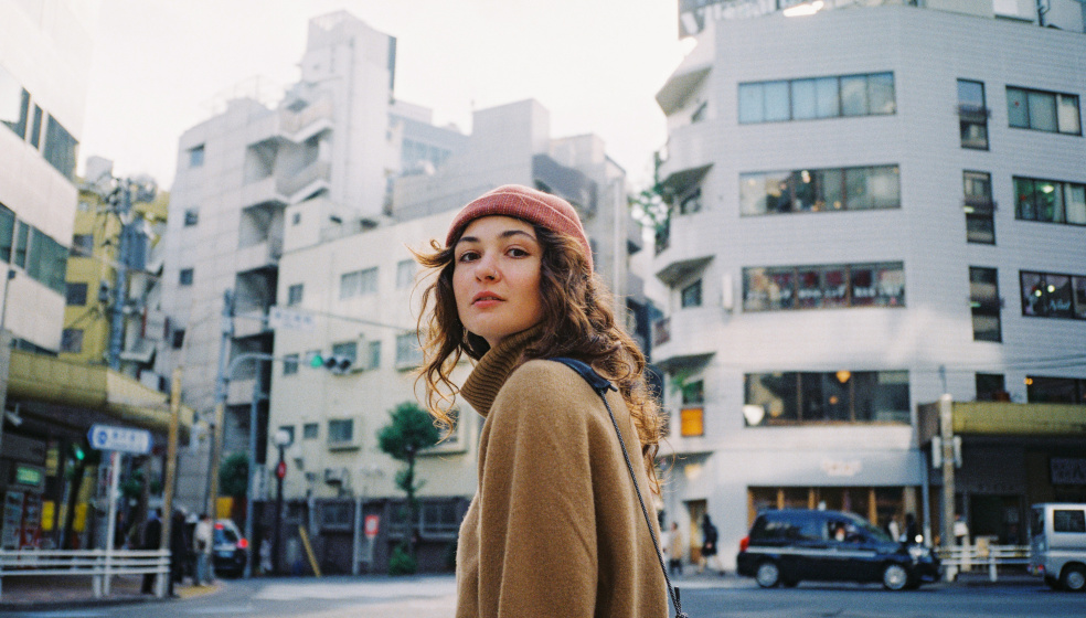 Eine Person mit langen Haaren und einer roten Mütze steht auf einer Straße in einer städtischen Umgebung, umgeben von mehreren mehrstöckigen Gebäuden und vorbeifahrenden Autos. Die Atmosphäre wirkt belebt und urban.